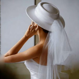 Puede incluir: Un sombrero blanco con una corona redondeada y ala ancha, combinado con un velo transparente y fluido. El sombrero es de diseño clásico, adecuado para bodas o eventos formales. El velo cae por la espalda, añadiendo un toque de elegancia.