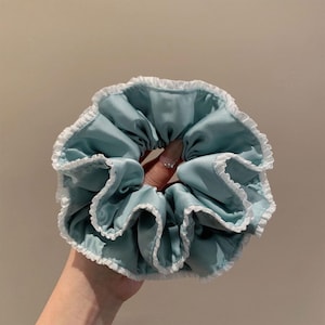 Peut inclure: Un chouchou en satin bleu clair avec une bordure en dentelle blanche. L'accessoire pour cheveux est froncé et froissé, créant un aspect volumineux. Le chouchou est tenu dans une main.