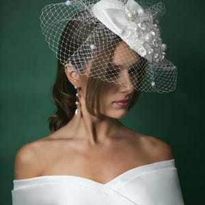 May include: A white bridal headpiece with a delicate net veil. The headpiece features a bow, pearls, and floral accents. The woman in the image is wearing a white off-the-shoulder dress and pearl earrings. The background is a dark green.
