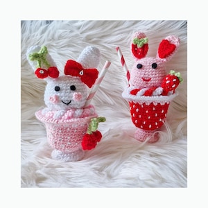 Puede incluir: Dos figuras de conejitos de ganchillo con forma de sundae. Una es blanca con un lazo rojo y detalles de cereza, la otra es rosa con detalles de fresa. Ambas tienen pajitas a rayas y están sobre un fondo blanco y esponjoso.