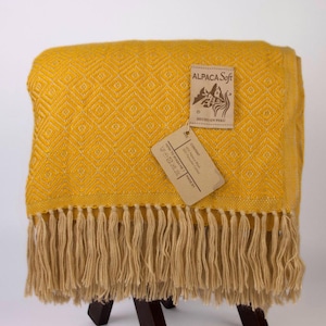 May include: A mustard yellow alpaca wool blanket with a diamond pattern and fringe. The blanket has a tag that reads "ALPACA Soft" and "Hecho en Peru". The blanket is draped over a dark brown wooden stool.