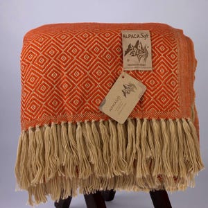 Könnte beinhalten: Eine orangefarbene und cremefarbene gewebte Decke mit einem Rautenmuster und Fransen. Die Decke ist über einen kleinen Hocker drapiert. Das Etikett lautet "ALPACA Soft" und "Hecho en Peru".