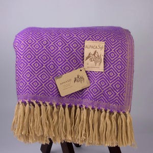 May include: A purple and tan woven alpaca blanket with a diamond pattern and tan fringe. The blanket has two tags that read "ALPACA Soft" and "HECHO EN PERU". The blanket is draped over a wooden stand.