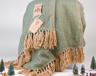 May include: A sage green woven throw blanket featuring a repeating diamond pattern and long beige fringe along the edges. Two rectangular paper tags are attached to the fabric, both printed with the text ALPACA Soft and a mountain graphic.