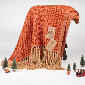 Op de afbeelding: Een geweven plaid in roestoranje en crème met een herhalend ruitpatroon en lange, geknoopte franjes aan de onderkant. Twee rechthoekige papieren labels met de tekst Alpaca Soft zijn met een wit touwtje aan de stof bevestigd.