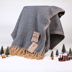 May include: A woven throw blanket featuring a dark blue and light gray geometric diamond pattern with beige fringe trim. Two rectangular paper tags are attached, one labeled Alpaca Soft and the other featuring a llama illustration.
