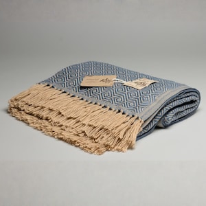 Peut inclure: Une couverture jetée tissée et pliée présentant un motif géométrique en losanges bleu et crème. La couverture est finie avec de longues franges de couleur crème sur le bord et comprend deux petites étiquettes en papier attachées par une ficelle blanche.