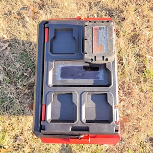 May include: A black and red tool organizer with multiple compartments and a battery pack. The organizer is rectangular and has a handle. The battery pack is black and gray with orange accents. The organizer is sitting on grass.