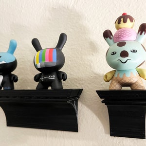 May include: Three colorful toy figurines on black shelves. The figurines are a blue and black bunny, a black bunny with a television screen for a face, and a light blue bunny with a brown and pink ice cream cone on its head.