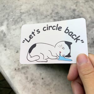 May include: A white sticker with a cartoon cat illustration. The cat is white with black spots and is crying a blue puddle. The text "Let's circle back" is above the cat.