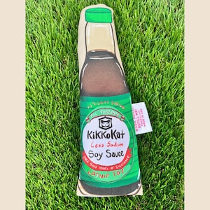May include: A catnip toy in the shape of a soy sauce bottle. The toy is green, brown, and beige, with the text "KikkoKat Less Sodium Soy Sauce" and "Catnip Toy". The toy is placed on green grass.