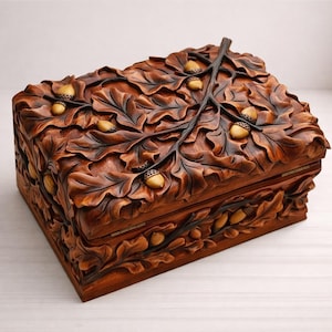 May include: A rectangular wooden box with a carved design of oak leaves and acorns. The box is stained in shades of brown and features a hinged lid. The acorns are a lighter shade of brown.