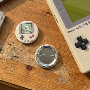 Op de afbeelding: Een crèmekleurige knoop met een retro videogame-ontwerp, met een klein scherm met een verdrietig gezicht en Japanse tekst. De knoop ligt naast een vintage Nintendo Game Boy op een houten oppervlak.