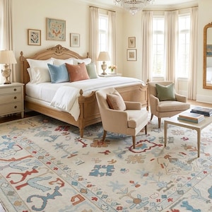 Op de afbeelding: Slaapkamer met een licht houten bedframe, wit beddengoed en sierkussens in blauw, roest en groen. Twee beige fauteuils en een vloerkleed met patronen met blauwe, rode en beige accenten. Een kroonluchter hangt aan het plafond.