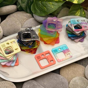 May include: A collection of colorful, square-shaped acrylic accessories in various colors, including pink, yellow, and blue. The accessories are stacked on a white marble tray, surrounded by smooth gray stones and green foliage.