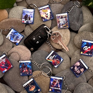 May include: A collection of miniature video game case keychains, featuring titles like "Spider-Man 2" and "Grand Theft Auto". The keychains are attached to silver key rings and are scattered on a bed of smooth, gray stones. A car key and house key are also present.