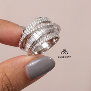 Peut inclure: Une bague en argent avec plusieurs bandes, certaines incrustées de petites pierres rondes et transparentes, d'autres de pierres rectangulaires et transparentes. La bague a un design moderne et entrelacé. La marque "LUXEERVA" est visible.
