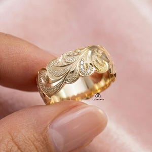 May include: A gold-colored ring with an intricate floral design. The ring features a textured surface with small, sparkling stones. The ring is held in a hand against a soft pink background. The ring is a band style.
