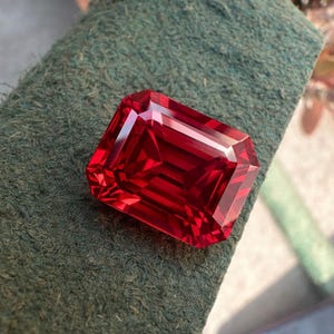 May include: A faceted, rectangular-cut, deep red gemstone. The gem has a brilliant, reflective surface, showcasing its internal structure and color. The stone is set against a textured, dark green background.