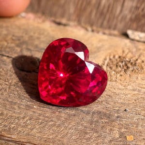 Puede incluir: Una gema de rubí facetada en forma de corazón. El rubí es de un color rojo intenso y vibrante, con muchas facetas que reflejan la luz. La gema está sobre una superficie de madera rústica.