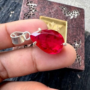May include: A silver pendant featuring a large, faceted, teardrop-shaped red gemstone. The gemstone is secured by small silver prongs and has a silver loop for a chain. The pendant is shown against a decorative background.
