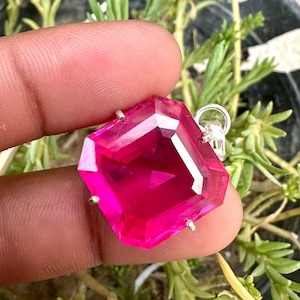 Puede incluir: Un colgante de piedra preciosa rosa intenso facetada con una fianza de plata. El colgante tiene forma rectangular con múltiples facetas, creando un efecto brillante. La piedra preciosa se sujeta con pequeñas garras de plata.