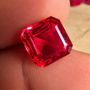 以下が含まれることがあります： 鮮やかな赤色の長方形にカットされた、ファセット加工された宝石。 宝石は、鮮やかな反射面とシャープなエッジとコーナーを持っています。 宝石は指の間に挟まれ、その色と透明度を示しています。 宝石の形状はエメラルドカットです。