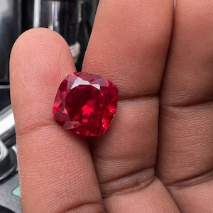 Könnte beinhalten: Ein facettierter, kissenförmiger, roter Edelstein, gehalten in einer Hand. Die Facetten des Steins fangen das Licht ein und zeigen seine Farbe und Klarheit. Der Stein hat eine quadratische Form mit abgerundeten Ecken und ähnelt einem Rubin.