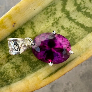 Könnte beinhalten: Ein silberner Anhänger mit einem ovalen, facettierten, tiefvioletten Edelstein. Der Anhänger hat eine dekorative Öse mit einem durchbrochenen Design. Der Edelstein wird von vier silbernen Zinken gehalten.