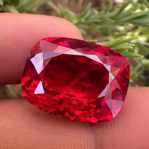 Peut inclure: Une pierre précieuse taillée à facettes, de forme coussin, d'un rouge vif. La gemme présente de nombreuses facettes qui réfléchissent la lumière, créant un effet scintillant. La pierre est tenue entre les doigts, avec un arrière-plan vert flou.
