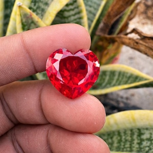 May include: A heart-shaped, red gemstone with multiple facets. The gem is held between fingers, highlighting its brilliant cut and vivid colour. The gemstone's facets reflect light, creating a sparkling effect. The background features green and yellow foliage.