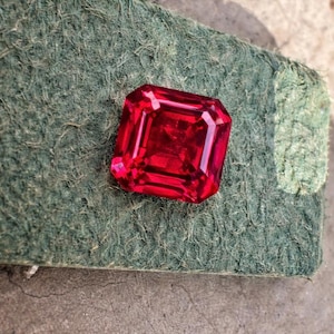 May include: A vibrant, faceted, red gemstone with an emerald cut. The gem is displayed on a textured, green surface. The gemstone's facets reflect light, creating a brilliant display of color.