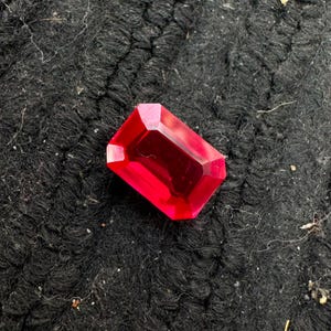 Può includere: Una gemma rosso brillante con taglio a smeraldo rettangolare, caratterizzata da bordi sfaccettati che riflettono la luce. La pietra presenta una tonalità cremisi profonda e satura e una superficie lucida e vitrea ben definita.