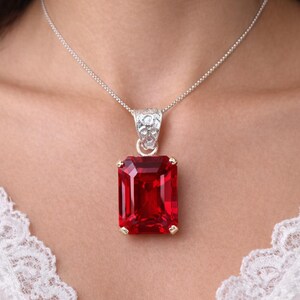 Può includere: Una collana d'argento con un grande pendente rettangolare di pietra rossa. Il pendente è incastonato in una cornice color oro e ha un'ansa d'argento strutturata. La collana è esposta su sfondo bianco.