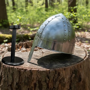 May include: A silver-colored medieval helmet with rivets, resting on a tree stump. A black display stand is to the left. The helmet has a pointed top and a face guard. The background is a blurred forest scene.