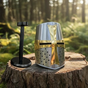 Puede incluir: Un casco medieval plateado y dorado con un diseño de cruz, exhibido sobre un tocón de madera. El casco tiene una construcción remachada y una cruz dorada en la parte delantera. Un soporte negro está al lado del casco.