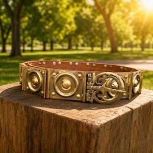 May include: A brown leather belt with ornate brass hardware. The belt features a series of rectangular brass plates with circular and geometric designs, connected by a brown leather strap. The buckle is a detailed brass design. The belt is displayed on a wooden block.