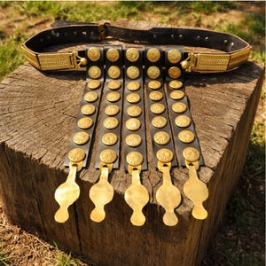 May include: A black leather belt with gold-colored decorative elements. The belt features a series of gold medallions and hanging pendants. The belt is displayed on a wooden surface.