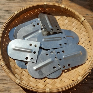 May include: A collection of silver-colored metal hardware pieces with rounded ends and multiple holes, arranged in a woven bamboo basket. Each piece has a rectangular section with holes and a curved, slightly raised section.
