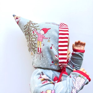 Op de afbeelding: Een baby draagt een blauwe en rode romper met capuchon met een kerstboom- en elfendesign. De capuchon heeft een rood-witte gestreepte rand.
