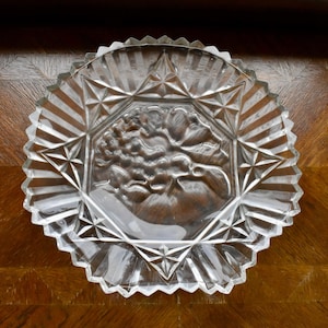 May include: Clear glass decorative plate with a starburst and geometric design. The center of the plate features a floral relief. The plate has a scalloped edge and is set on a wooden surface.