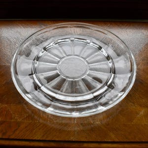 May include: A clear glass plate with a circular design. The plate has a raised rim and a textured center. The plate is sitting on a wooden surface with a geometric pattern. The plate is likely for serving food.