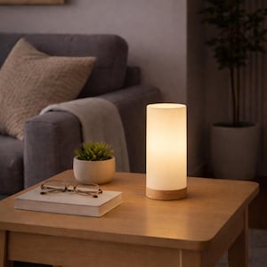 May include: A cylindrical table lamp with a frosted white glass shade and a light-toned wooden base. The lamp emits a warm, soft glow and is positioned on a wooden table next to a closed book, a pair of reading glasses, and a small potted succulent.