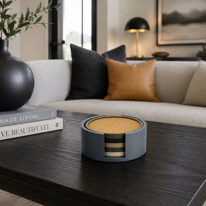 May include: A set of circular coasters with cork tops and layered, multi-colored bases, housed in a matching grey cylindrical holder. The coasters feature a stackable design with alternating bands of dark grey, tan, and cream colors visible on the sides.