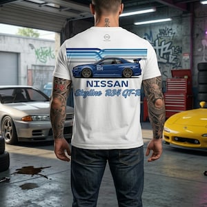 May include: White t-shirt featuring a blue Nissan Skyline R34 GT-R car graphic. The design includes the car's silhouette, the text "NISSAN Skyline R34 GT-R", and blue stripes. The person is wearing blue jeans.