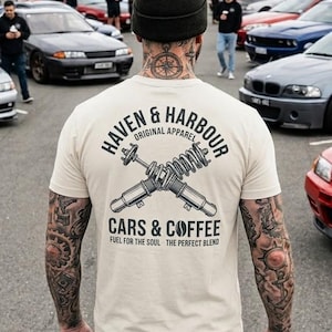 May include: A beige t-shirt with the text "HAVEN & HARBOUR ORIGINAL APPAREL" and "CARS & COFFEE FUEL FOR THE SOUL THE PERFECT BLEND" printed on the back. The design includes crossed shock absorbers. The person is wearing a black beanie and blue jeans.