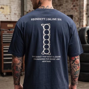 May include: A navy blue short-sleeve t-shirt featuring a white graphic of an inline six engine gasket on the back. The text reads RB26DETT | INLINE SIX, THE GASKET THAT BUILT A LEGEND, ENGINEEERED FOR BOOST JDM HERITAGE in white lettering.