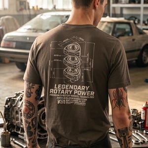 Könnte beinhalten: Braunes T-Shirt mit weißer Grafik eines Rotationsmotors und dem Text "LEGENDARY ROTARY POWER." Die Person trägt das Shirt in einer Garage. Autoteile und Werkzeuge sind im Hintergrund zu sehen.