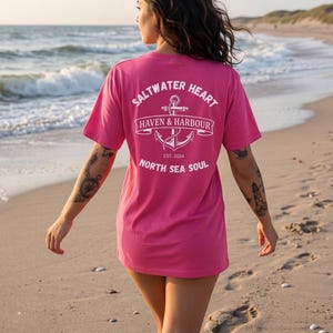 May include: A bright pink t-shirt with the text "Saltwater Heart Haven & Harbour North Sea Soul" and an anchor graphic on the back. The shirt is worn on a beach with the ocean in the background.