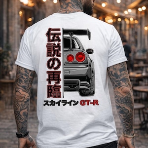 Puede incluir: Camiseta blanca con un gráfico de un coche deportivo gris con luces traseras rojas y un alerón trasero. El texto japonés está impreso verticalmente en el lado izquierdo y el texto "スカイライン GT-R" está impreso en la parte inferior.